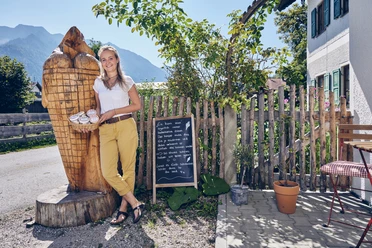 Bio-Hof-Eiswerkstatt Person steht lächelnd mit Eiskorb neben einer Holzskulptur auf einem Hof bei Sonnenschein.