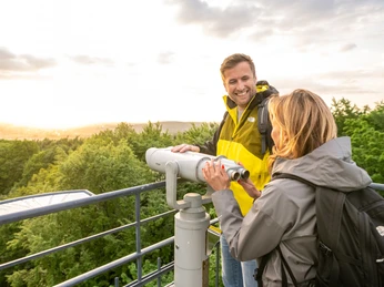 Zwei Menschen stehen lächelnd bei einem Fernrohr auf einem Aussichtsturm, mit Blick auf bewaldete Hügel.