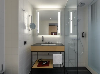 Modernes Badezimmer mit beleuchtetem Spiegel, Waschbecken, Duschkabine und Badzubehör auf dunklem Boden.