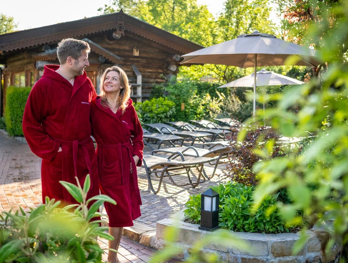 Herford-2025-034-©Teutoburger Wald - Pro Herford GmbH - D. Ketz.jpg Ein Paar in roten Bademänteln spaziert lächelnd durch eine grüne Gartenlandschaft mit Liegestühlen.