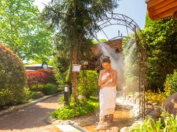 Eine Frau steht in einem Dampfbadgarten unter einem dekorativen Metallbogen, umgeben von grüner Vegetation.