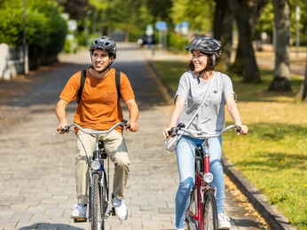 herford-promenade-teutoburger-wald-tourismus-h-tornow-008-jpg.jpg Zwei Personen radeln bei sonnigem Wetter auf einem gepflasterten Weg durch eine grüne, parkähnliche Umgebung.