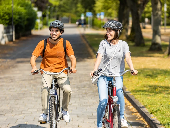 herford-promenade-teutoburger-wald-tourismus-h-tornow-008-jpg.jpg Zwei Personen radeln bei sonnigem Wetter auf einem gepflasterten Weg durch eine grüne, parkähnliche Umgebung.