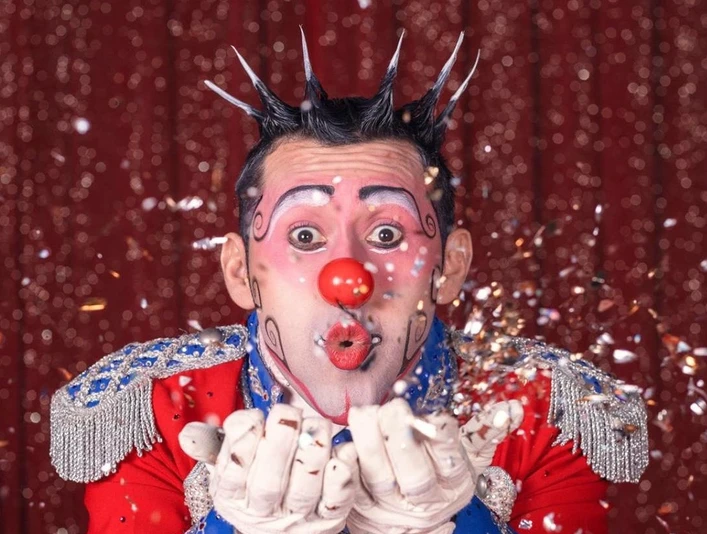 Great Christmas Circus Frankfurt - Henry the prince of clowns Clown in bunter Uniform pustet glitzernden Konfetti vor rotem Vorhang.Clown in a colorful uniform blows glittering confetti in front of a red curtain.