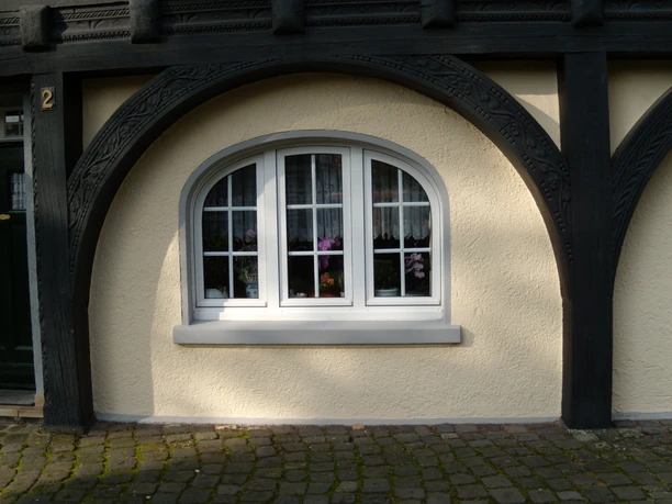 Fachwerkfassade mit dekorativem Rundbogen und weiß gerahmtem Fenster, florale Dekoration im Inneren.