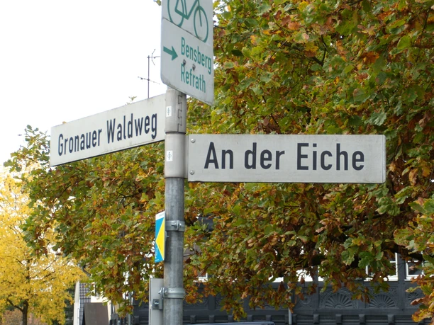 Straßenschilder mit den Namen "Gronauer Waldweg" und "An der Eiche" vor Herbstbäumen.