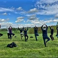 Yogawanderung Menschen praktizieren Yoga in einer malerischen Landschaft. Sie stehen auf einem grünen Feld unter blauem Himmel.