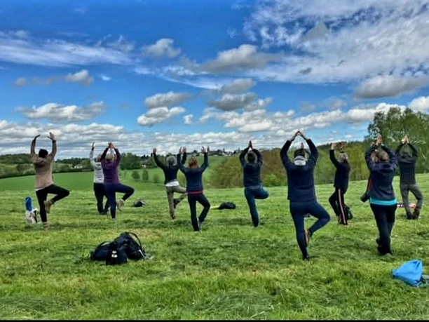 Yogawanderung Menschen praktizieren Yoga in einer malerischen Landschaft. Sie stehen auf einem grünen Feld unter blauem Himmel.