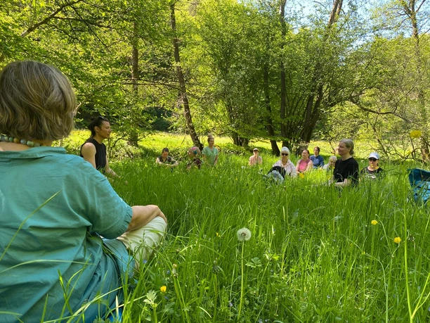 Yogawanderung Personengruppe sitzt entspannt im grünen Gras eines sonnigen, von Bäumen umgebenen Parks.