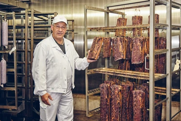 Metzgerei Haller Mitarbeiter in weißem Kittel zeigt geräuchertes Fleisch in einer Metzgerei.
