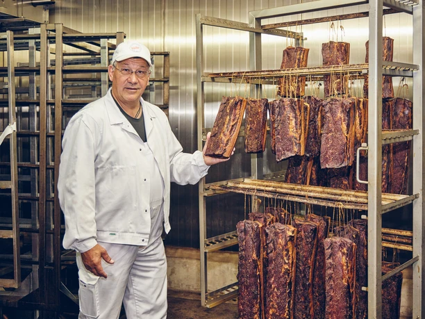 Metzgerei Haller Mitarbeiter in weißem Kittel zeigt geräuchertes Fleisch in einer Metzgerei.