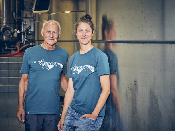 Brauerei Karg Zwei Personen in blauen T-Shirts mit Bergmotiv stehen vor Brauerei-Tanks.
