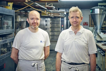 Bäckerei Josef Krätz Zwei Männer in Arbeitskleidung stehen in einer Backstube vor Öfen.
