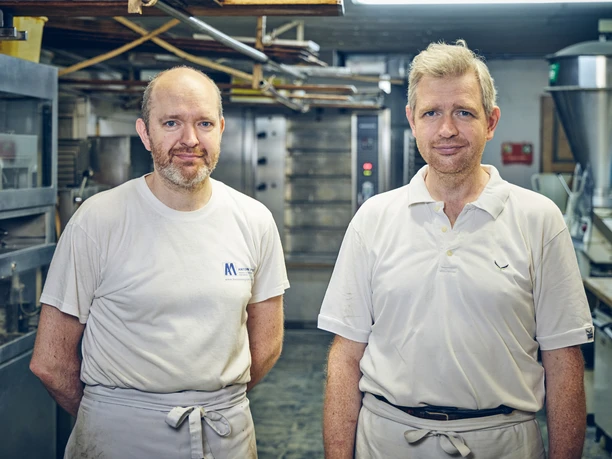 Bäckerei Josef Krätz Zwei Männer in Arbeitskleidung stehen in einer Backstube vor Öfen.