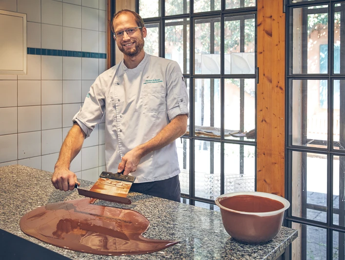 Schokoladenmanufaktur Barbara Krönner Mann in weißem Kochhemd temperiert Schokolade auf einer Arbeitsplatte in heller Küche.