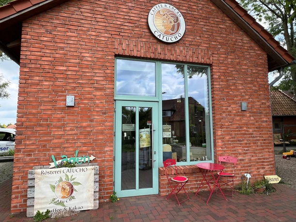 Roastery with brick building, large glass front, turquoise door and two red chairs in front of it.