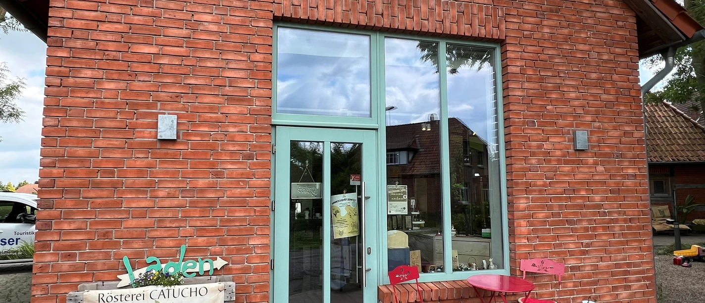 Catucho Rösterei mit Backsteingebäude, großer Glasfront, türkisfarbiger Tür und zwei roten Stühlen davor.