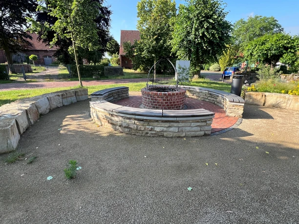 Halbrunder, mit Steinen eingefasster Sitzbereich mit einem zentralen, steinernen Brunnen in sonniger Umgebung.