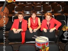 Drei Personen mit roten Westen und Hemden sitzen fröhlich auf Theaterstühlen.