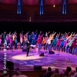 Ein großer Chor in bunter Kleidung tritt in einer beleuchteten Konzerthalle auf, dirigiert von einer Person.
