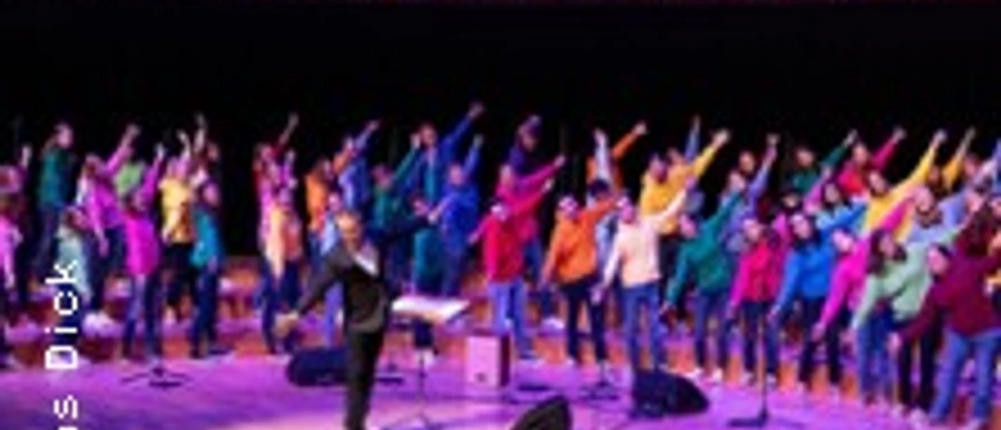 Ein großer Chor in bunter Kleidung tritt in einer beleuchteten Konzerthalle auf, dirigiert von einer Person.