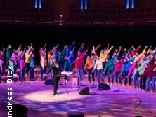 Ein großer Chor in bunter Kleidung tritt in einer beleuchteten Konzerthalle auf, dirigiert von einer Person.