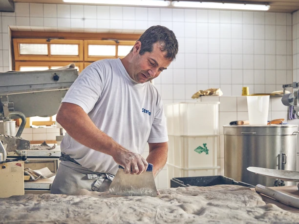 Bäckerei Luidl Bäcker in Arbeitskleidung knetet Teig in heller, gefliester Backstube.