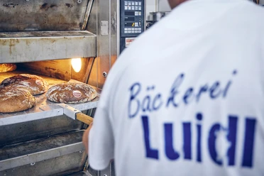 Bäckerei Luidl Bäcker in Luidl-T-Shirt nimmt frisch gebackenes Brot aus dem Ofen.