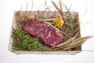 Murnau-Werdenfelser-Rind Ein Stück rohes Rindfleisch liegt auf Stroh, umgeben von frischen Kräutern und Getreide.