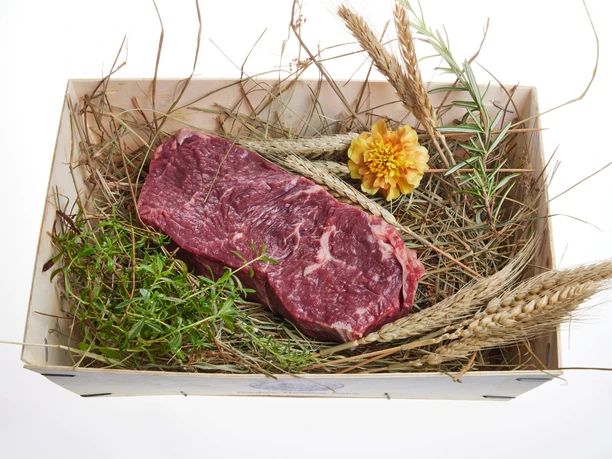 Murnau-Werdenfelser-Rind Ein Stück rohes Rindfleisch liegt auf Stroh, umgeben von frischen Kräutern und Getreide.