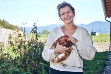 Promberger Hof Eine lächelnde Frau hält ein Huhn, umgeben von einem blühenden Garten.