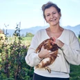 Promberger Hof Eine lächelnde Frau hält ein Huhn, umgeben von einem blühenden Garten.