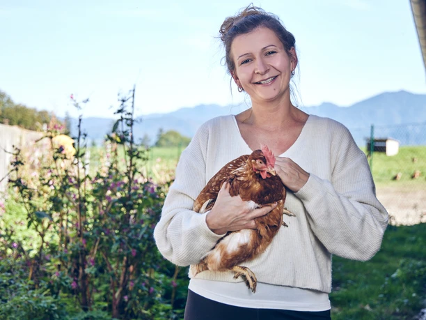 Promberger Hof Eine lächelnde Frau hält ein Huhn, umgeben von einem blühenden Garten.