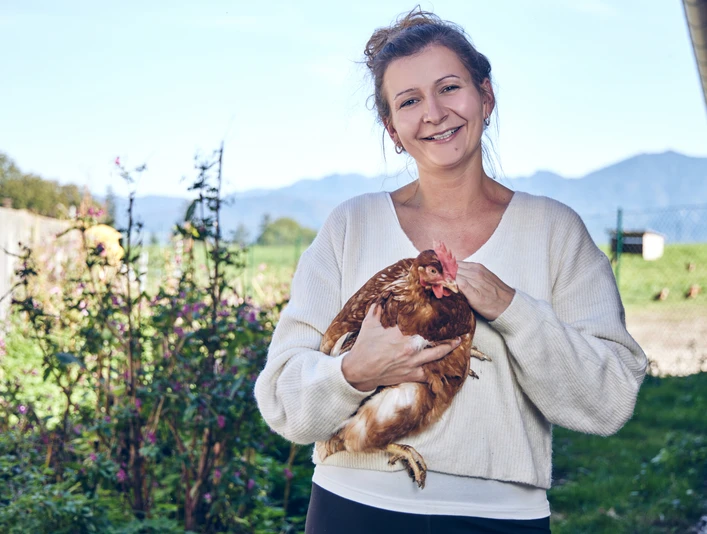 Promberger Hof Eine lächelnde Frau hält ein Huhn, umgeben von einem blühenden Garten.