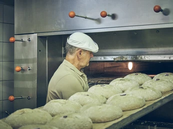 Bäckerei Sand Bäcker legt Brotlaibe in einen Ofen; links Metallgriffe, Fokus auf Brotteig.