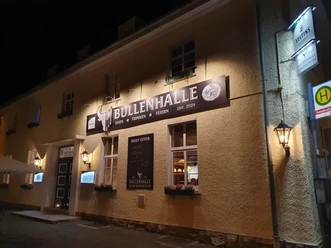 Eine Außenaufnahme der Bullenhalle bei Nacht, beleuchtet, einladendes lokales Restaurant mit nostalgischem Flair.