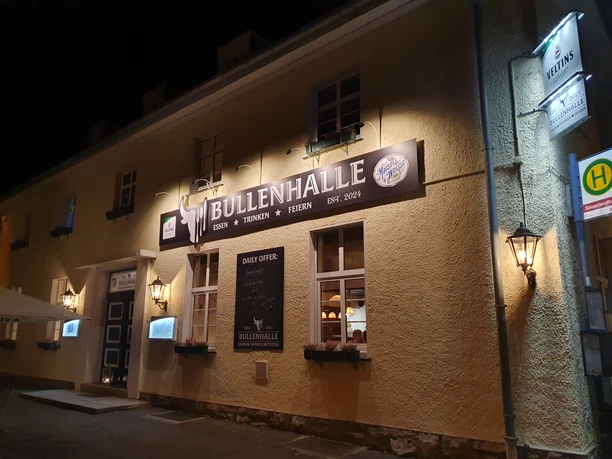 Eine Außenaufnahme der Bullenhalle bei Nacht, beleuchtet, einladendes lokales Restaurant mit nostalgischem Flair.