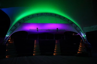 Open Air Kino ralf-pauli Nächtlich beleuchtetes Open-Air-Amphitheater. Die geschwungene Überdachung erstrahlt in leuchtenden Grün- und Lilatönen und verleiht dem Ort eine einladende Atmosphäre. Die Sitzreihen sind ebenfalls beleuchtet und führen den Blick zur Bühne.