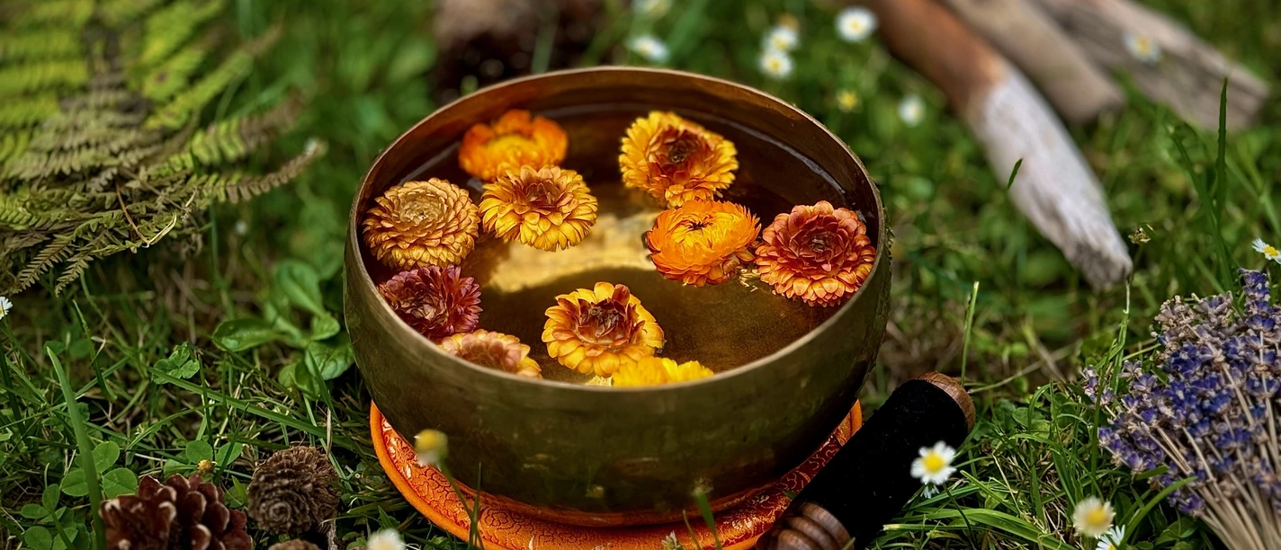 Achtsame Klang- & Naturauszeit Goldene Klangschale mit schwimmenden Blumenblüten, umgeben von Naturdekoration auf einer Wiese.