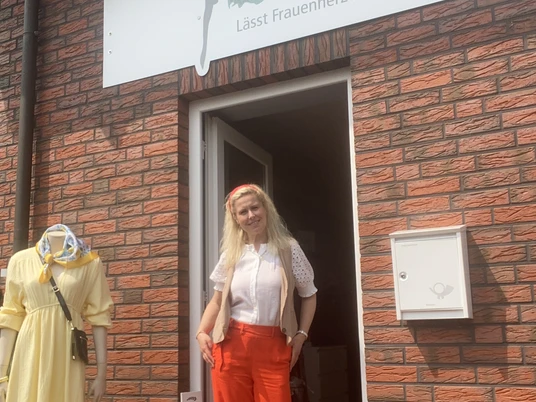 cindys-frauenzimmer.JPG Eine Frau mit langen blonden Haaren steht lächelnd in einem farbenfrohen Outfit in der Tür eines Ladens mit der Aufschrift "Cindys Frauenzimmer".