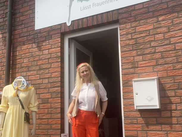 cindys-frauenzimmer.JPG Eine Frau mit langen blonden Haaren steht lächelnd in einem farbenfrohen Outfit in der Tür eines Ladens mit der Aufschrift "Cindys Frauenzimmer".