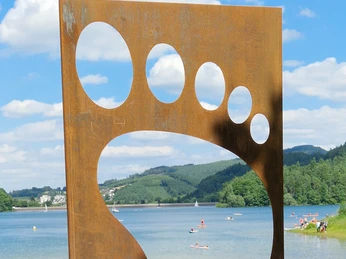 Barfusspfad Hennesee Eingang Eine große, metallene Fußskulptur vor dem Hennesee, Badegäste im Gras liegend.