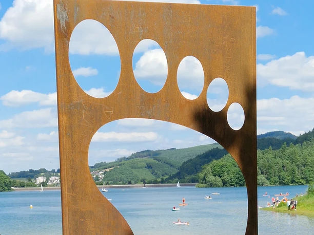 Barfusspfad Hennesee Eingang Eine große, metallene Fußskulptur vor dem Hennesee, Badegäste im Gras liegend.