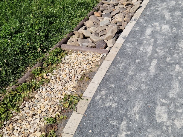 Barfusspfad Hennesee Kiesel Das Bild zeigt einen Barfußpfad mit verschiedenen Texturen: Gras, Kieselsteine und größere Steine. Der Pfad windet sich in einer sanften Kurve. Im Hintergrund sind grüne Pflanzen sichtbar.