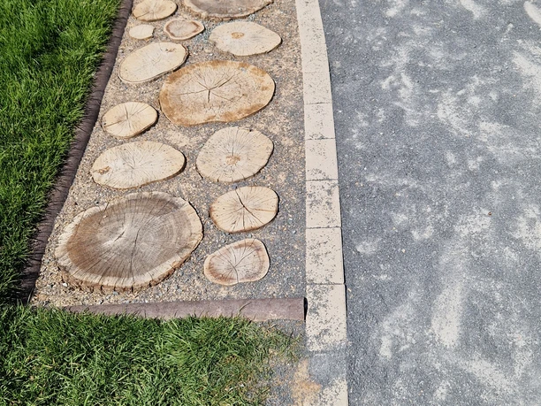 Barfusspfad Hennesee Baumscheiben Das Bild zeigt einen Teil des Barfußpfades am Hennesee. Auf der linken Seite sind große runde Holzscheiben in den Boden eingelassen, die zum Barfußgehen einladen. Rechts daneben verläuft ein glatter gepflasterter Weg mit einer hellen Begrenzung aus Beton oder Stein. Auf beiden Seiten des Weges sind grüne Grasflächen zu sehen.