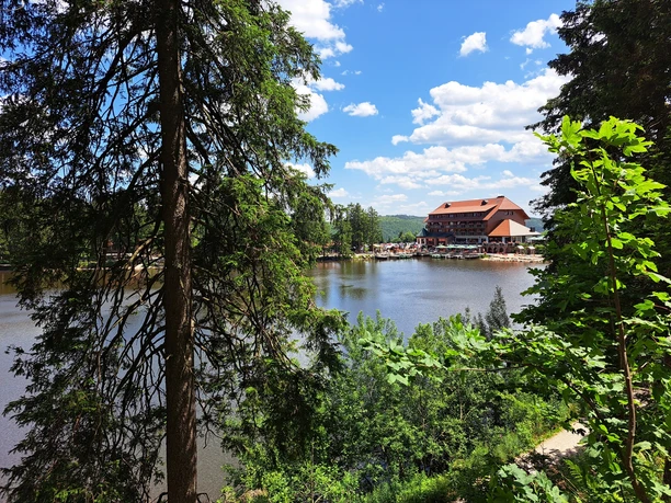 Blick durch Bäume auf den Mummelsee mit Berghotel vor blauem Himmel mit Wolken
