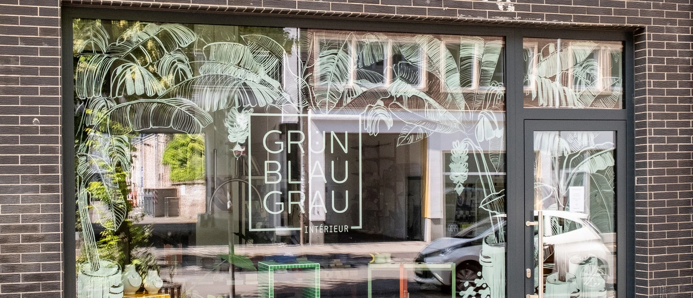 GrünBlauGrau Intérieur The façade of a store with large windows shows the lettering "GrünBlauGrau Intérieur" in front of a decorated shop window.