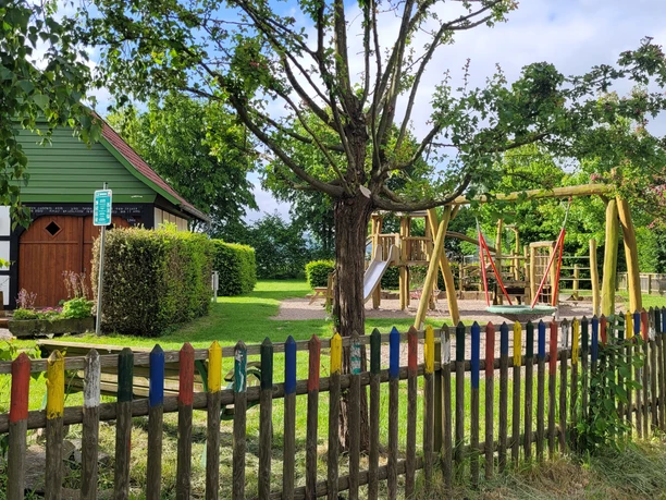 Ortsteil Stockausen Lübbecke Ein kleiner Spielplatz mit Klettergerüst und bunter Umzäunung, umgeben von Bäumen an einem sonnigen Tag.