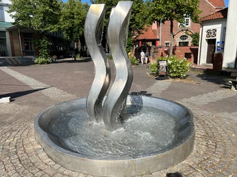 Drei elegant geschwungene Edelstahlrohre formen einen Brunnen, umgeben von Blumen und Backsteinbauten.