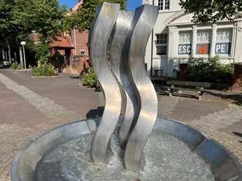 Statue Die 3 Grazien Drei geschwungene Metallsäulen erheben sich aus einem Wasserbrunnen vor einem historischen Gebäude.